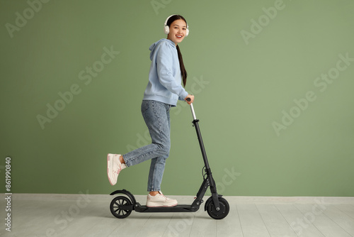 Beautiful young Asian woman in headphones riding modern electric kick scooter near green wall