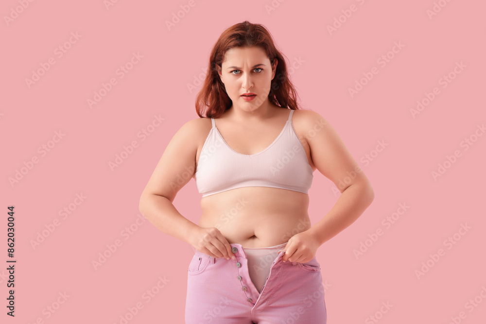 Overweight young worried woman in tight jeans on pink background. Weight gain concept Overweight young worried woman in tight jeans on pink background. Weight gain concept