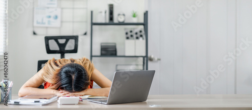 Tired woman resting her head on a desk in a modern office setting, with a laptop and shelves in the background.