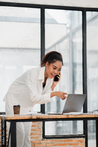 Confident young woman multitasking in a contemporary office, using a laptop and smartphone for business communication.