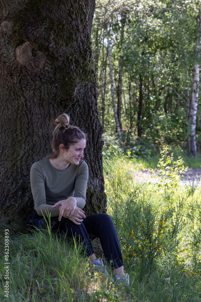 A young woman is peacefully seated by a tree in a beautiful forest, cherishing the serene and natural surroundings A young woman is peacefully seated by a tree in a beautiful forest, cherishing the serene and natural surroundings