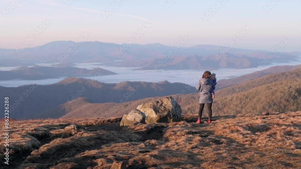 A mother and child stand on a rocky mountain peak at sunrise, admiring the expansive, serene landscape of rolling hills and distant valleys. High quality 4k footage A mother and child stand on a rocky mountain peak at sunrise, admiring the expansive, serene landscape of rolling hills and distant valleys. High quality 4k footage