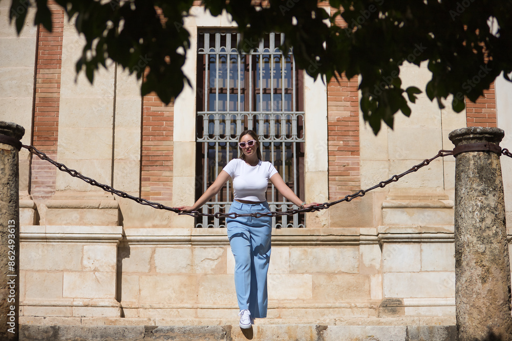 Young beautiful woman with brown hair, sunglasses, white t-shirt and blue pants, posing leaning on a huge chain between two columns while visiting the city. Concept beauty, fashion, trend. Young beautiful woman with brown hair, sunglasses, white t-shirt and blue pants, posing leaning on a huge chain between two columns while visiting the city. Concept beauty, fashion, trend.