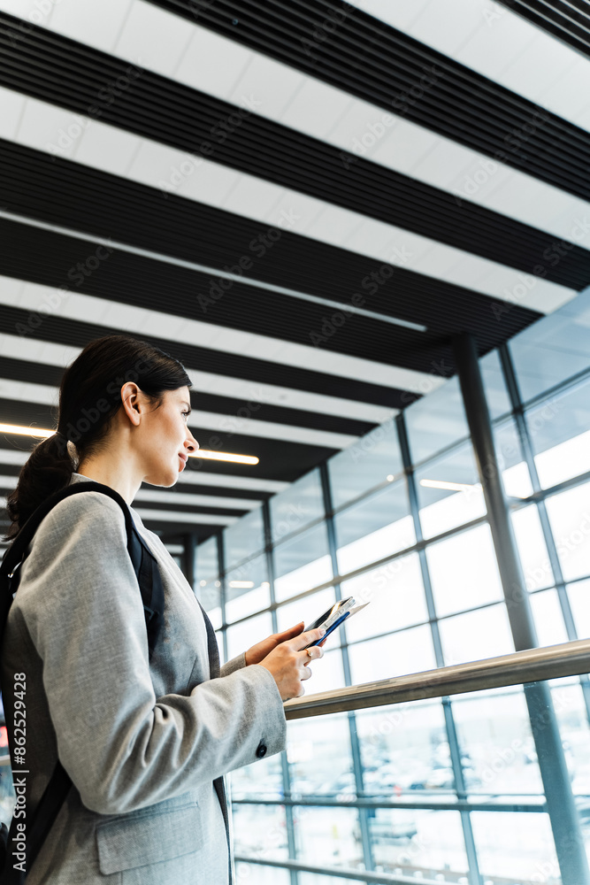 Tourist with smartphone, documents and backpack in the airport lounge is looking through the panoramic window. Smiling caucasian woman waiting for flight to start vacation in 2024. Tourist with smartphone, documents and backpack in the airport lounge is looking through the panoramic window. Smiling caucasian woman waiting for flight to start vacation in 2024.