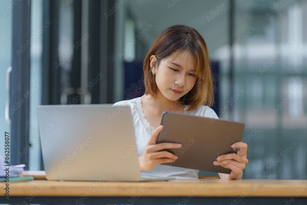 Young Asian women hand-holding and using digital tablets, work on laptop computers with books on the desk at home. Young Asian women hand-holding and using digital tablets, work on laptop computers with books on the desk at home.