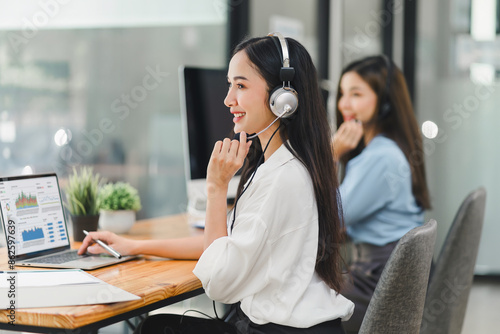 Smiling call center agents wearing headsets, working at desks with computers and documents in a modern office. Soft focus
