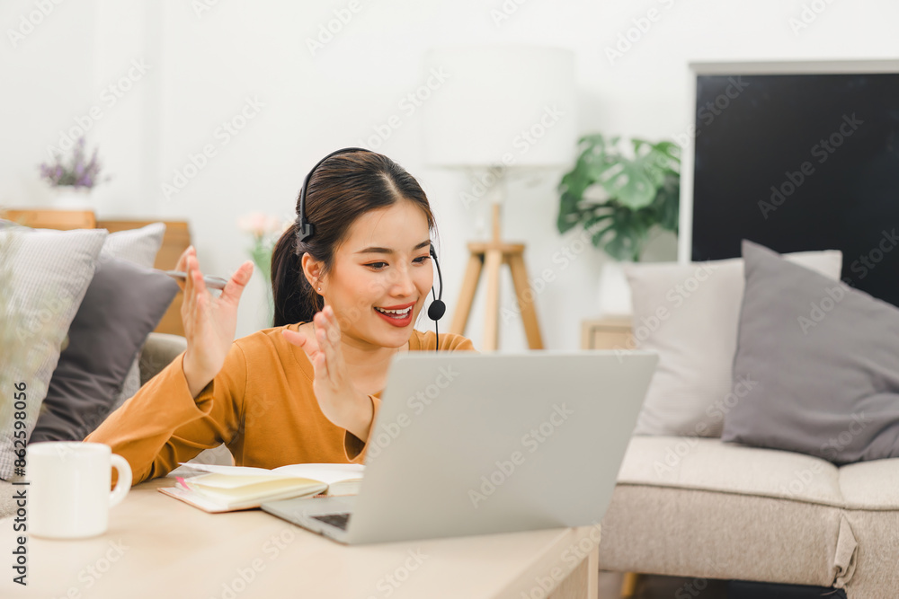 Smiling woman wearing a headset, engaged in video call while working from home with laptop and taking notes. Smiling woman wearing a headset, engaged in video call while working from home with laptop and taking notes.