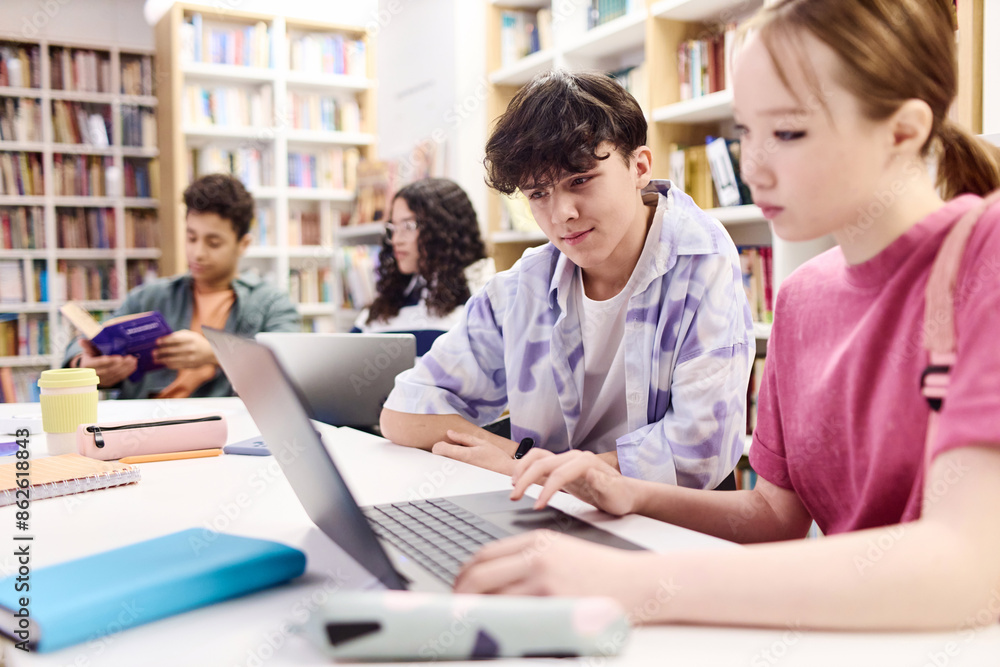 Portrait of young teenage boy looking at laptop screen in school library with group of students copy space Portrait of young teenage boy looking at laptop screen in school library with group of students copy space