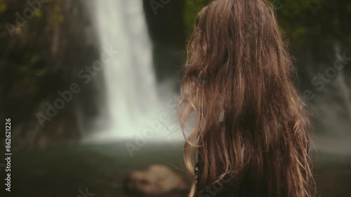 Beautiful woman standing against powerful Bali waterfall. Travel, tourism, holiday, freedom and active lifestyle concept. Indonesia, Bali island, Air Terjun Munduk, Melanting Waterfalls. Slow motion photo