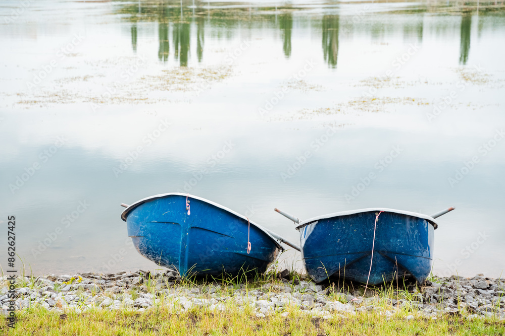 Two serene blue boats peacefully rest at the calm lakes shore, creating a picturesque scene surrounded by the beauty and tranquility of nature. The watercraft enhance the natural landscape Two serene blue boats peacefully rest at the calm lakes shore, creating a picturesque scene surrounded by the beauty and tranquility of nature. The watercraft enhance the natural landscape