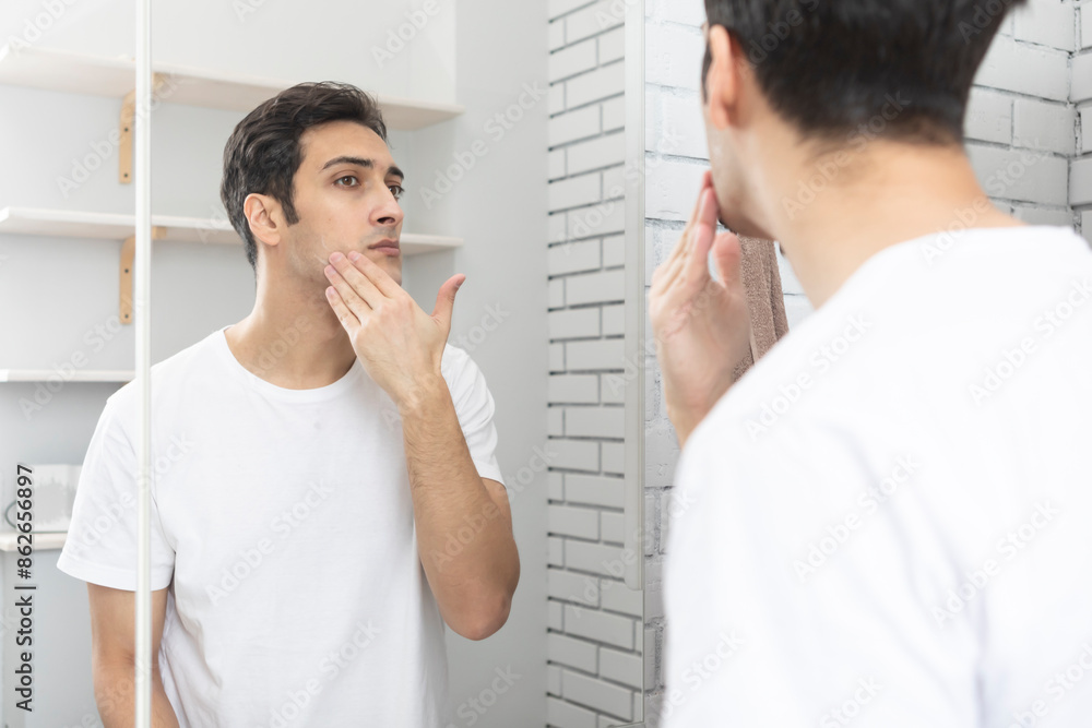 Handsome man looking at mirror and applying moisturizing cream on cheeks in bathroom. Groomed young guy doing skincare morning routine after taking a shower Handsome man looking at mirror and applying moisturizing cream on cheeks in bathroom. Groomed young guy doing skincare morning routine after taking a shower