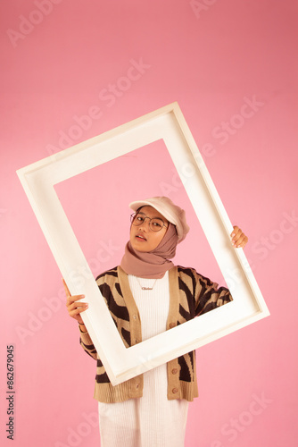 portrait of asian teenage woman posing with white frame and wearing hijab, white hat and glasses. pink background
