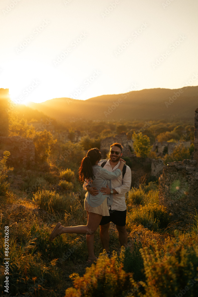 A couple embracing at sunset in a scenic mountain landscape. The warm sunlight highlights the greenery and ruins around them, creating a romantic and serene atmosphere. A couple embracing at sunset in a scenic mountain landscape. The warm sunlight highlights the greenery and ruins around them, creating a romantic and serene atmosphere.