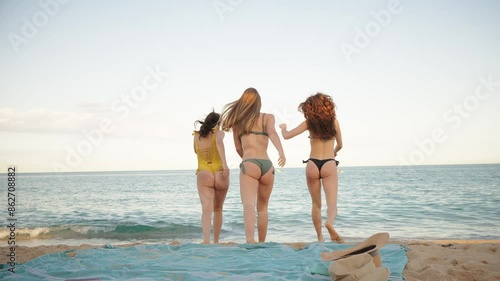 Rear view of three beauty young women spreading their arms on the sand of beach. Group of only girls in bikinis swimsuit enjoying the coast freely, looking out toward the sea. Summer vacations