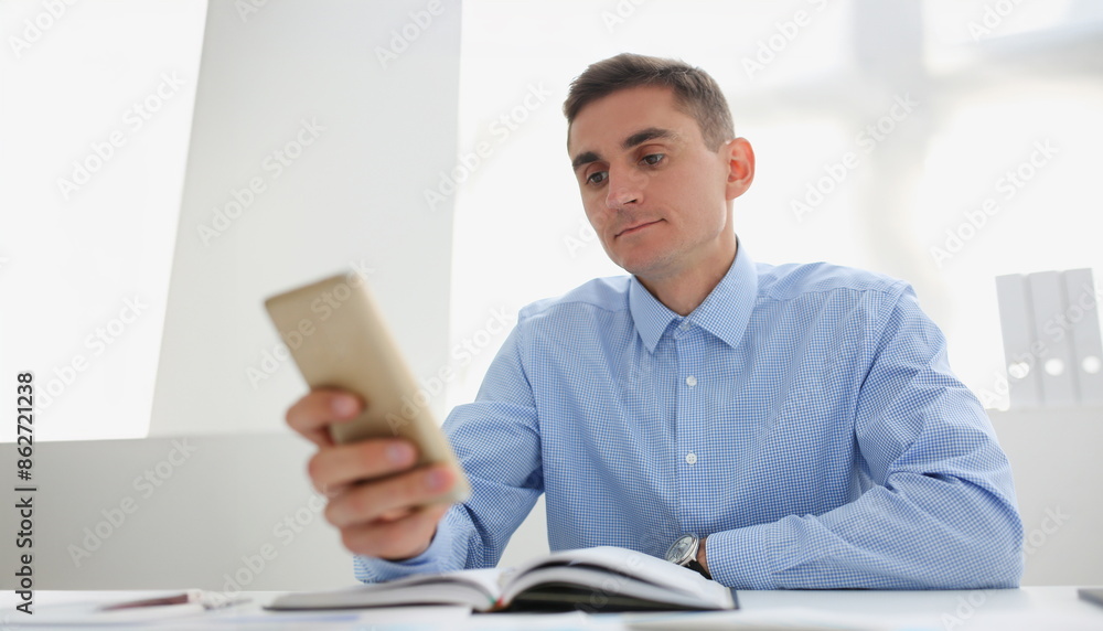 Young man in blue shirt looks at smartphone screen while sitting at his desk. Smartphone apps for planning working day concept Young man in blue shirt looks at smartphone screen while sitting at his desk. Smartphone apps for planning working day concept