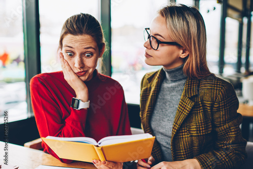 Shocked skilled student reading textbook with confused information during private lesson with qualified coach in coworking.Wondered hipster girl analyzing studying material together with tutor © BullRun