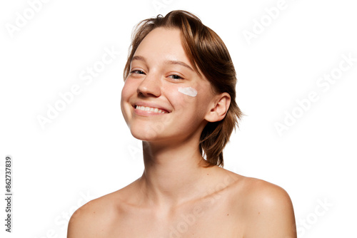 Well-kept woman with short brown hair smiles brightly, applying lotion to her cheek against white studio background. Concept of youth, beauty and fashion, cosmetic product, dermatology. Ad