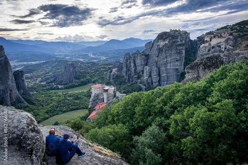 Stunning view from above of the monastery castles in Meteora, built on the tops of the rocks. Two tourists sit on a rock with their backs to the camera and watch the stunning Meteora sunset.
Kalambaka photo