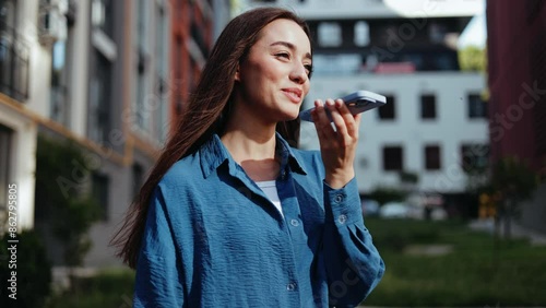 A young woman walks down an urban street and talks on her smartphone using the speakerphone feature.