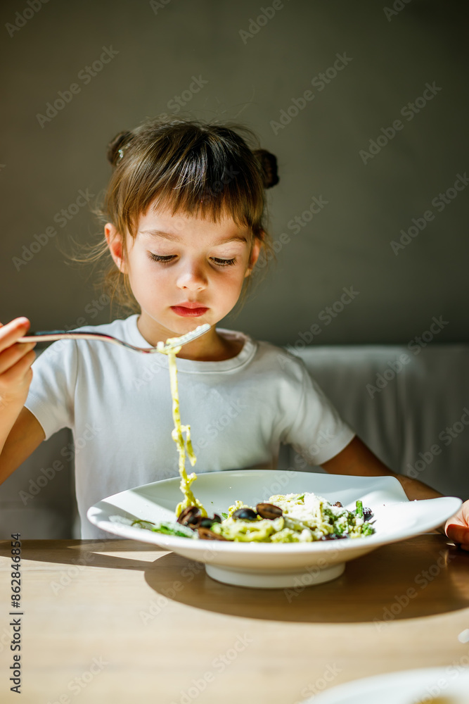 A young girl twisting pasta on a fork, eating a bowl of vegetarian pasta with green pesto and black olives