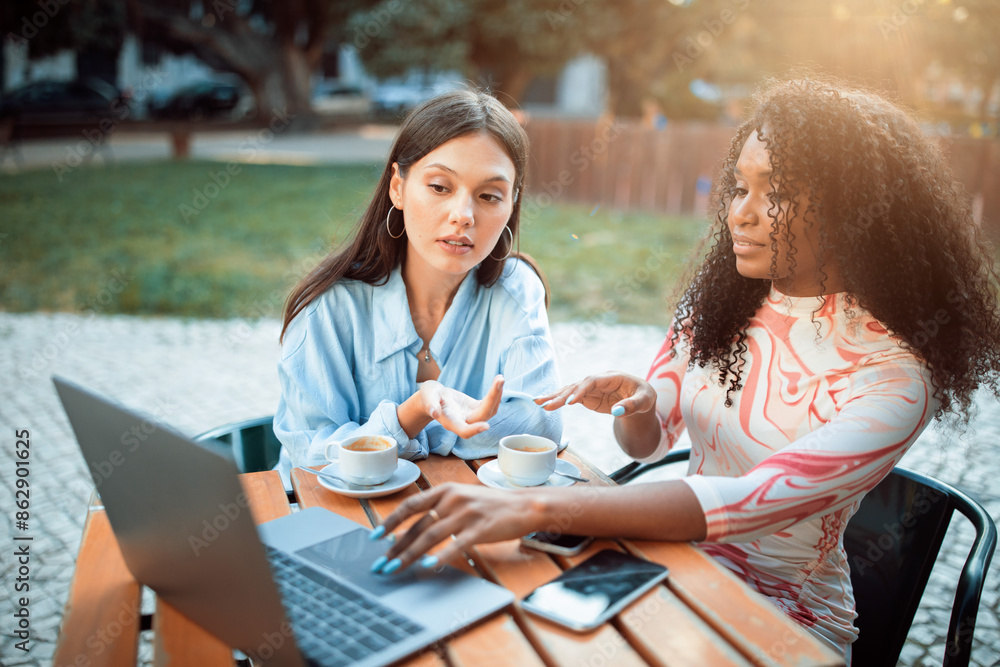 Two women are seated at a table outside a cafe. One woman is using a laptop computer while the other woman gestures and looks on. There is a cell phone on the table next to the laptop. Two women are seated at a table outside a cafe. One woman is using a laptop computer while the other woman gestures and looks on. There is a cell phone on the table next to the laptop.