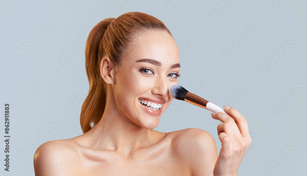 Fototapeta premium A woman with long blonde hair pulled back in a ponytail is applying makeup to her face with a brush. She is smiling and looking at the camera. The background is a light blue color.
