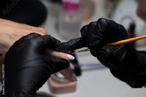 Close-up of a nail technician wearing black gloves, applying intricate nail art to a client's nails with a fine brush. The focus is on the detailed work and precision in the nail design. © Liana