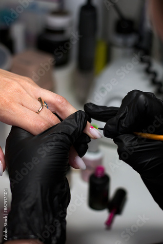 A professional manicurist in black gloves carefully applying intricate nail art to a client's nails, highlighting detailed design and precision in a salon setting. © Liana