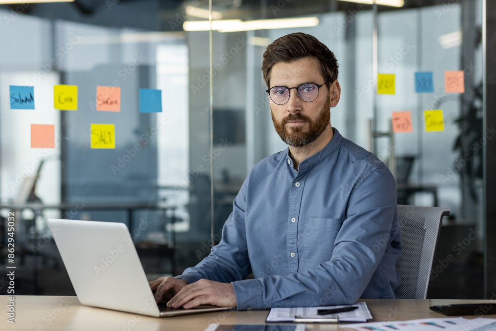 Portrait of a young self-confident and serious young businessman sitting in a modern office at a desk, using and working on a laptop, looking at the camera Portrait of a young self-confident and serious young businessman sitting in a modern office at a desk, using and working on a laptop, looking at the camera