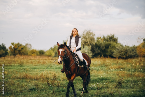 Foto Young beautiful blonde woman jockey rides a brown horse in a meadow at sunset in summer