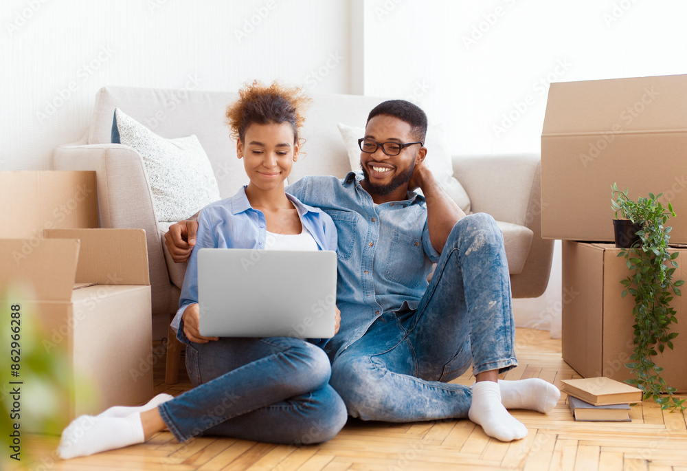 Relocation Concept. Spouses Browsing On Laptop Ordering Furniture For New Apartment Sitting On Floor Among Moving Boxes Indoor. Selective Focus Relocation Concept. Spouses Browsing On Laptop Ordering Furniture For New Apartment Sitting On Floor Among Moving Boxes Indoor. Selective Focus