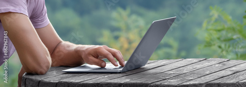 Hands of man using laptop computer and Internet on wooden table outdoor on summer vacation among nature. Working, reading news, studying, chatting remotely online. Alternative office workplace Banner