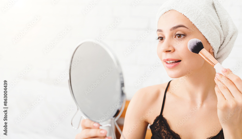 A woman is using a mirror to apply makeup with a brush in a sunny room. She is wearing a bathrobe and a towel on her head, suggesting its part of her morning routine. A woman is using a mirror to apply makeup with a brush in a sunny room. She is wearing a bathrobe and a towel on her head, suggesting its part of her morning routine.