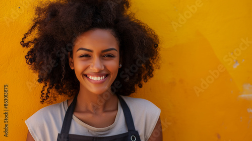 Alegre mulher afro-americana com cabelos cacheados, vestindo um avental, sorri contra uma parede amarela. photo
