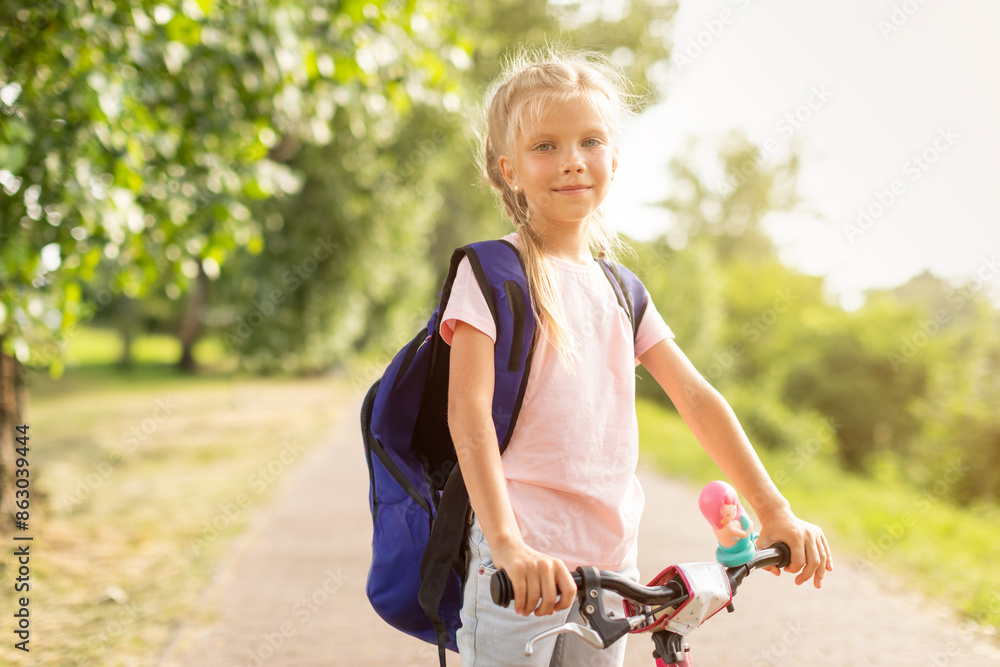 This image shows a young girl wearing a pink shirt and a blue backpack. She is riding a bike on a paved path in a park. The girl is looking at the camera and smiling. This image shows a young girl wearing a pink shirt and a blue backpack. She is riding a bike on a paved path in a park. The girl is looking at the camera and smiling.