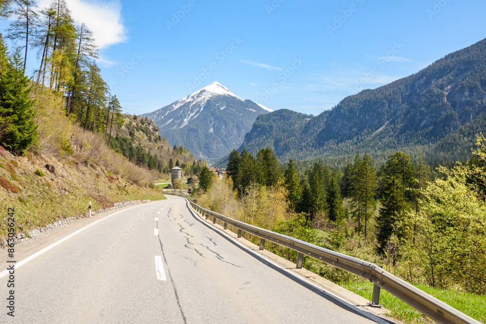 Fototapeta premium Empty winding mountain road through a forested valley in Switzerland on a clear spring day. Agricultural buildings along the road are visible in distance.