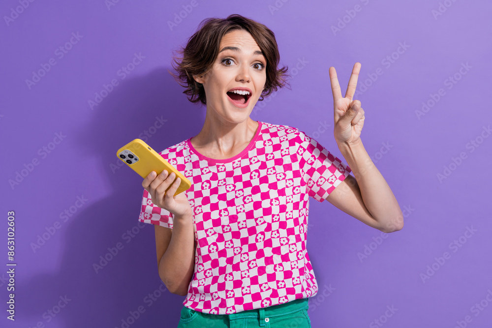 Photo of cheerful cute girl wear stylish clothes hold phone show v-sign special seasonal discount isolated on purple color background Photo of cheerful cute girl wear stylish clothes hold phone show v-sign special seasonal discount isolated on purple color background