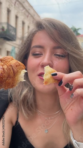 Vertical close up street video. Young adult joyful blonde white woman took piece from crispy croissant in city street cafe. Concept of birthday, holiday, cuisine, healthy eating, food. Cute face