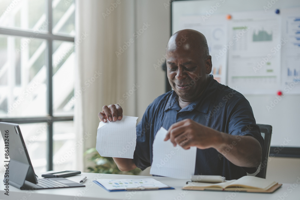 Businessman tearing up paper in frustration at his desk in a modern office space with a computer and charts in the background. Businessman tearing up paper in frustration at his desk in a modern office space with a computer and charts in the background.