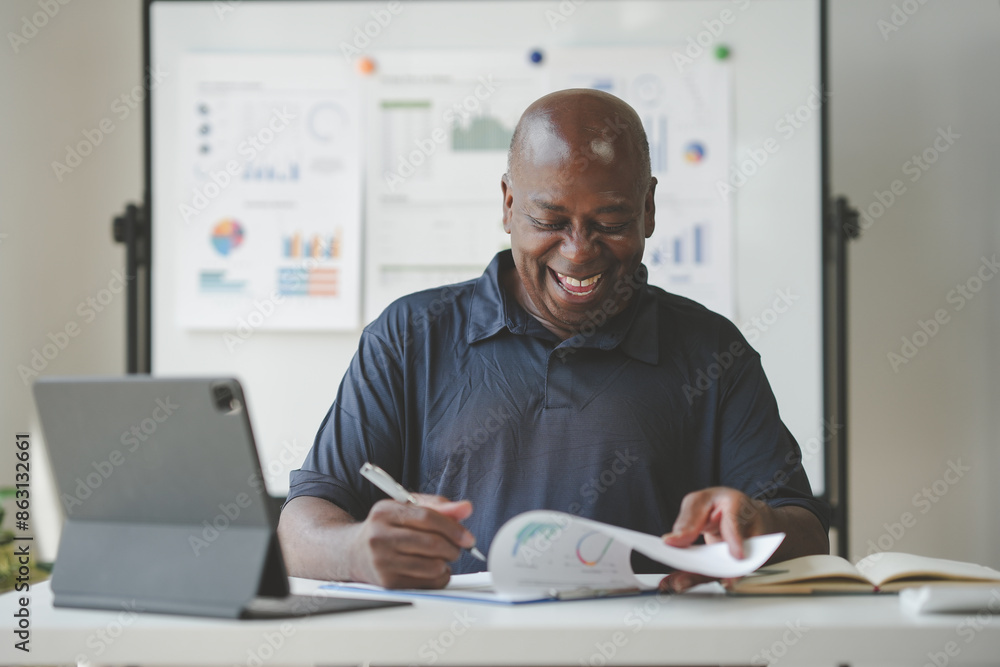 Smiling man in an office setting working on documents with a laptop, charts, and graphs in the background. He's focused and enjoying his task. Smiling man in an office setting working on documents with a laptop, charts, and graphs in the background. He's focused and enjoying his task.