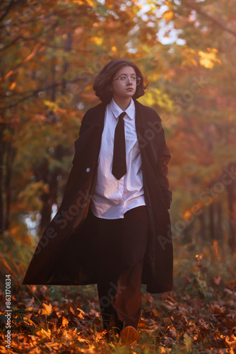 teenage girl in glasses and black raincoat walks in alley of autumn city park on background of autumn maples, autumn toning, autumn background, selective focus