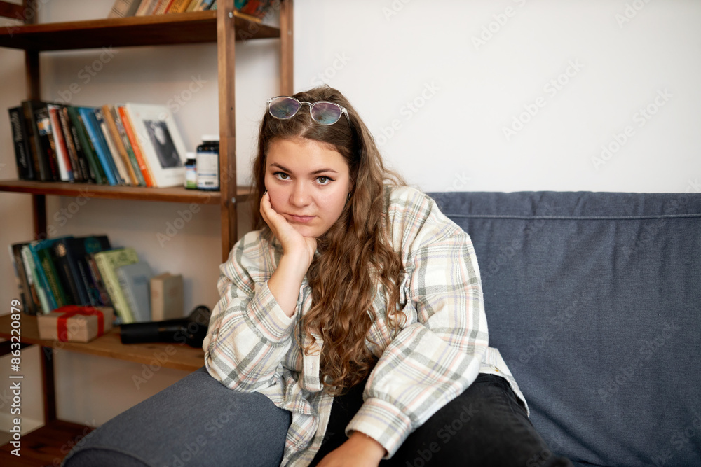 Tired plus size female in casual clothes sitting on couch holding her chin with hand, looking at camera with exhausted facial expression, having rest after finishing household chores Tired plus size female in casual clothes sitting on couch holding her chin with hand, looking at camera with exhausted facial expression, having rest after finishing household chores