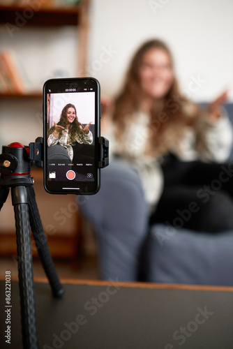 Selective focus on camera of smartphone standing on tripod recording podcast of female blogger or influencer gesticulating and sharing advice with subscribers or followers of her blog or vlog