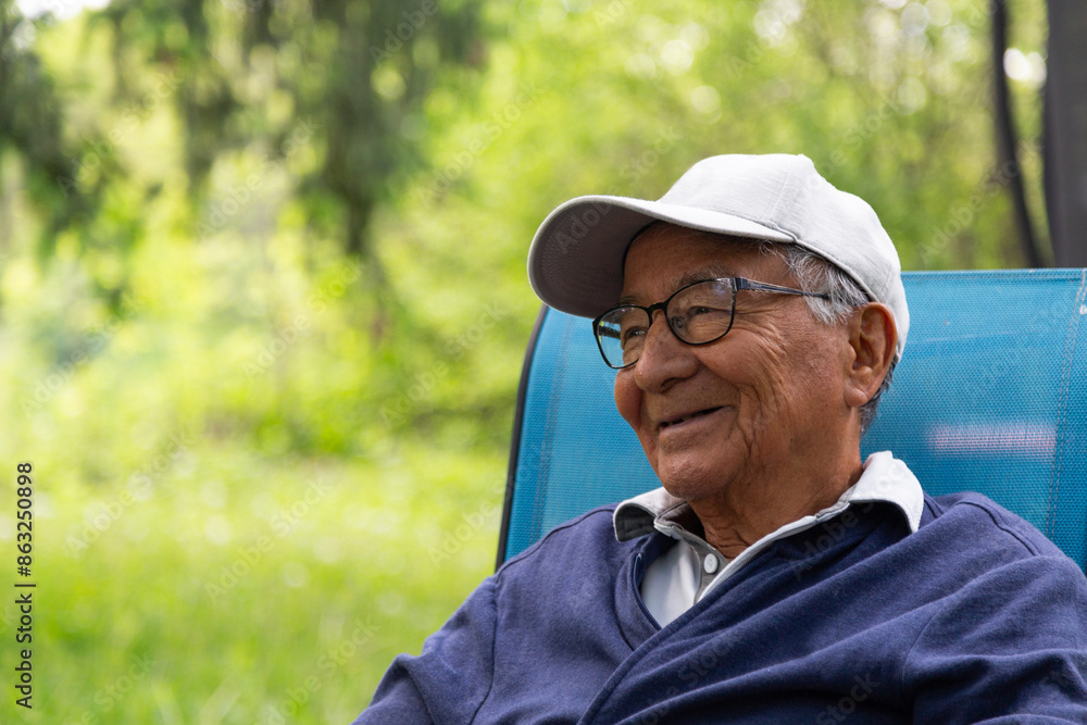 Fototapeta premium A man in a blue sweatshirt and white cap smiles. He is sitting on a chair in a park