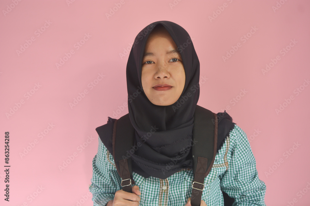 Portrait of a young Asian Muslim schoolgirl wearing a green shirt and black hijab while carrying a black bag on her back, showing enthusiastic expression and hand gesture Portrait of a young Asian Muslim schoolgirl wearing a green shirt and black hijab while carrying a black bag on her back, showing enthusiastic expression and hand gesture