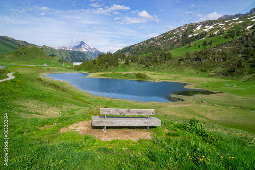 Panorama with a park bench on a hiking trail with a view of a lake and the surrounding mountains in the Arlberg area, snow-covered cloudy mountain peaks and green, flower-filled meadows in spring