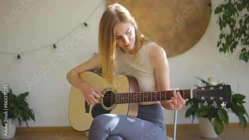 A serene young woman focused on playing acoustic guitar indoors, embodying dedication and passion for musicmaking in a peaceful and relaxing atmosphere with plants surrounding her photo