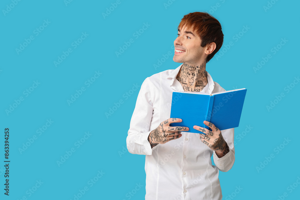 Happy handsome young tattooed man with book on blue background Happy handsome young tattooed man with book on blue background