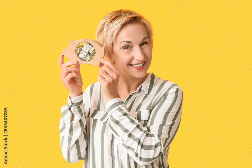 Beautiful mature woman with wooden piggy bank on yellow background Beautiful mature woman with wooden piggy bank on yellow background