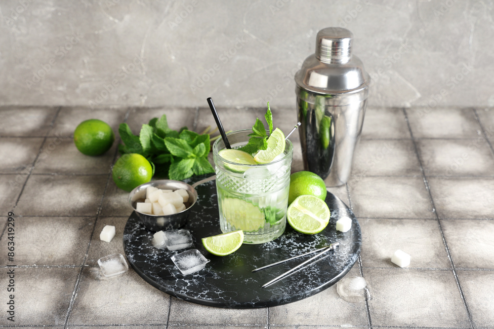 Glass of fresh Mojito cocktail with limes and shaker on grey tile table Glass of fresh Mojito cocktail with limes and shaker on grey tile table